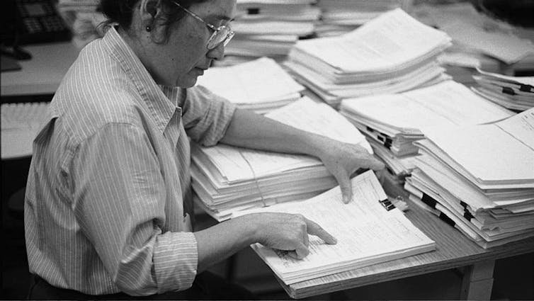 Person wearing glasses reviewing a stack of papers, surrounded by other stacks of papers on a desk