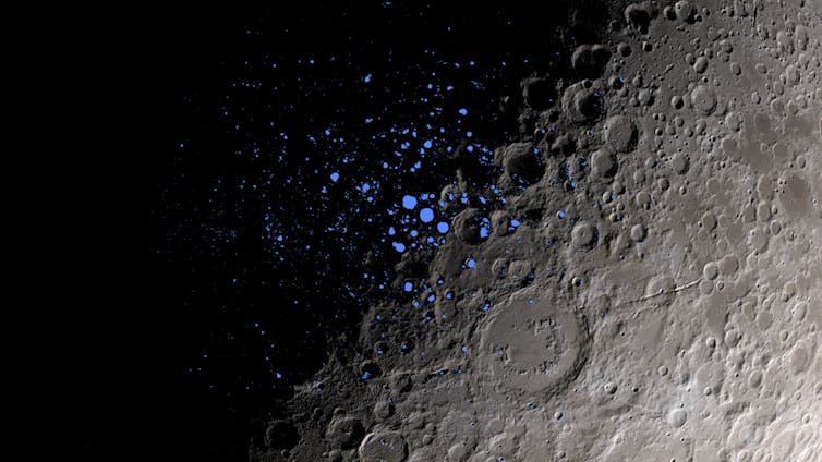 file-20231113-25-275gqy A close-up shot of the Moon's surface, with the left half covered in shadow, and the right half visible, with gray craters. Tiny blue dots in the center indicate PSRs.
